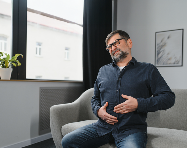 Ein Mann mittleren Alters sitzt mit schmerzverzerrtem Gesicht auf dem Sofa und hält sich den Bauch – typisches Anzeichen für Bauchschmerzen als mögliches Symptom von Darmkrebs.