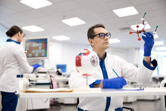 Ein Mann mittleren Alters hält im Vordergrund einen Aminosäuren-Bausatz hoch und macht sich Notizen, während im Hintergrund eine Frau vor einem Computer im Labor steht. Beide tragen blaue Handschuhe und weiße Kittel.