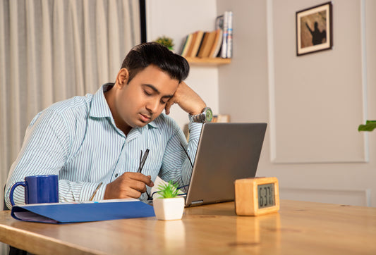 Ein Mann sitzt vor seinem Laptop ermüdet an einem Tisch. Auf dem Tisch steht noch sein blauer Becher, eine Blaue Arbeitsmappe, eine kleine Tischpflanze und ein Wecker 